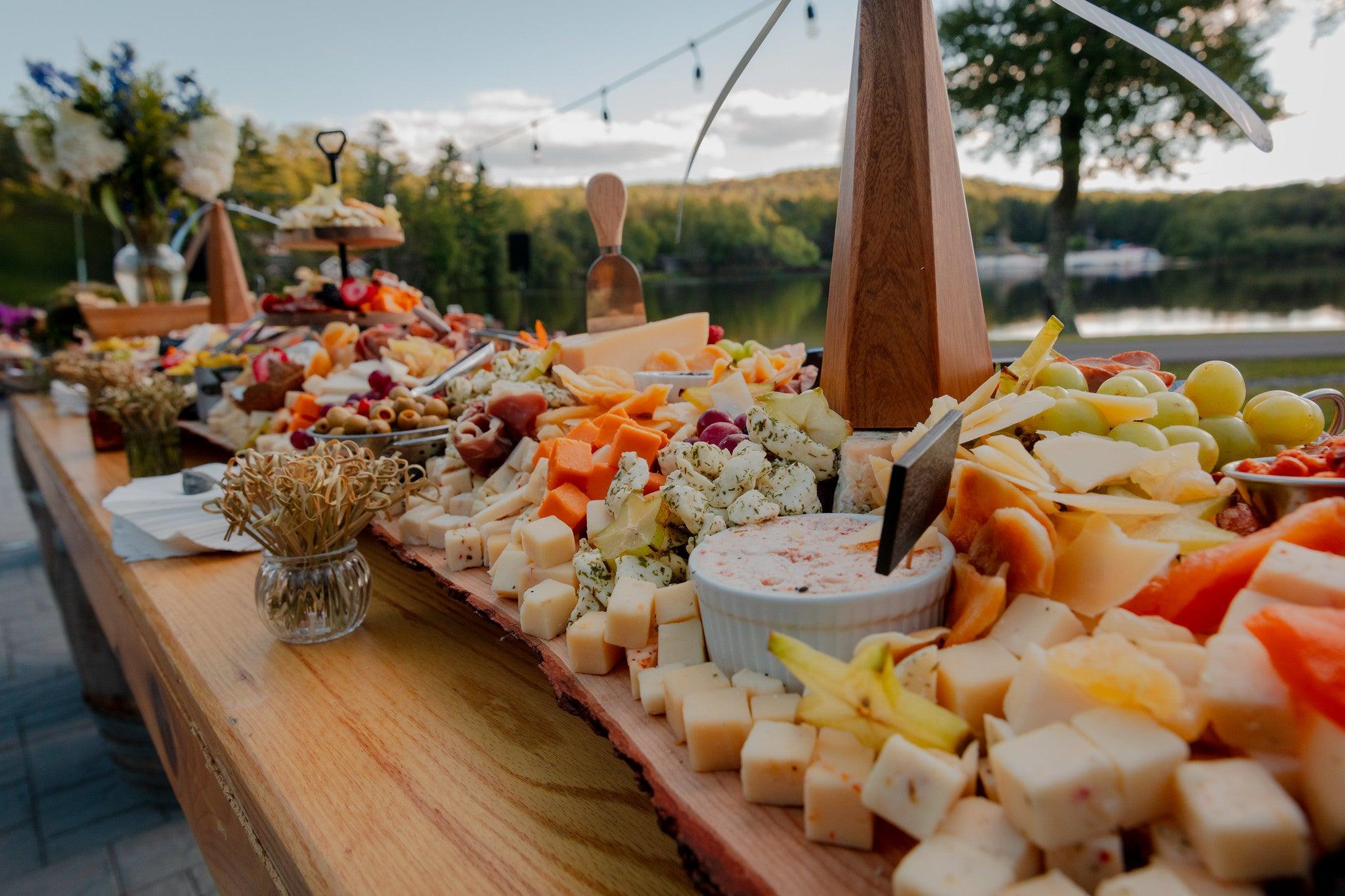 A long charcuterie board with cheese, fruits, meats, dips, and toothpicks on a wooden table in an outdoor setting.