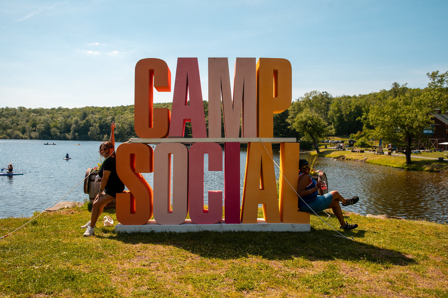 Large CAMP SOCIAL block sign in front of a lake with two women sitting on the sign